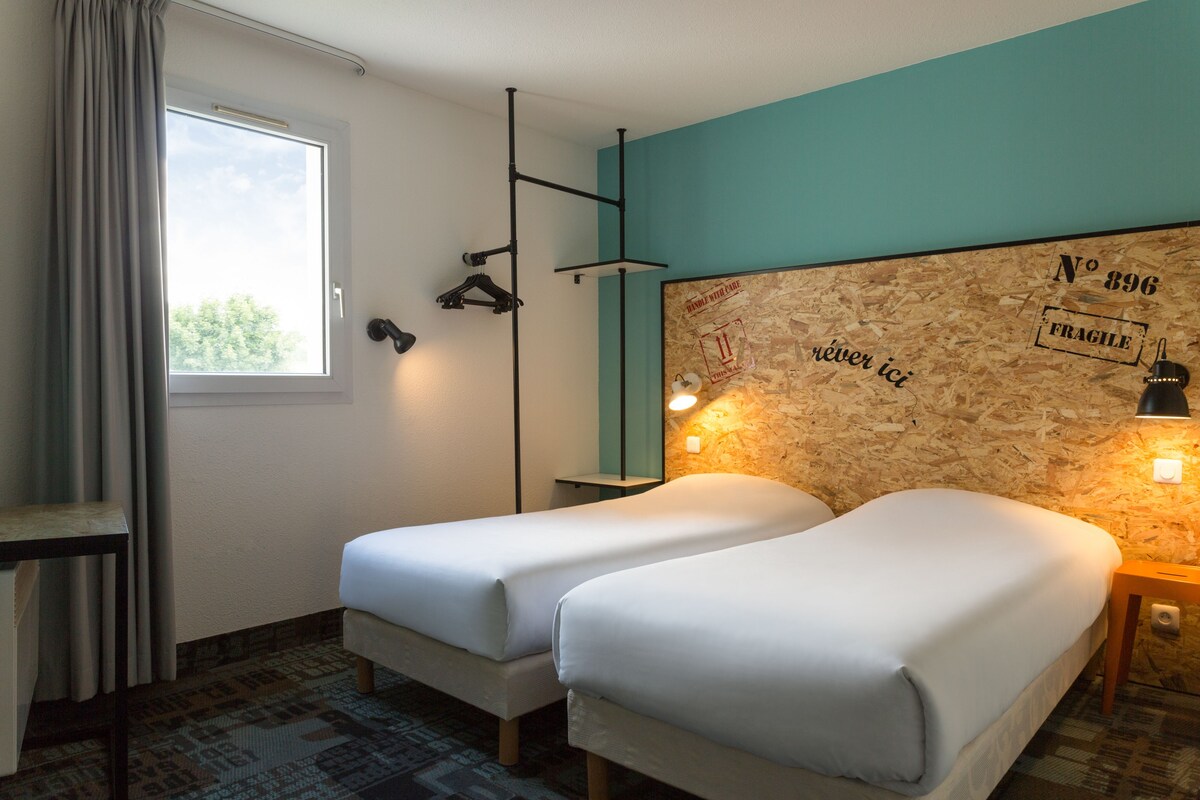 A room with two single beds, a window with curtains, a wall-mounted light, and a wooden textured wall with a number and word.