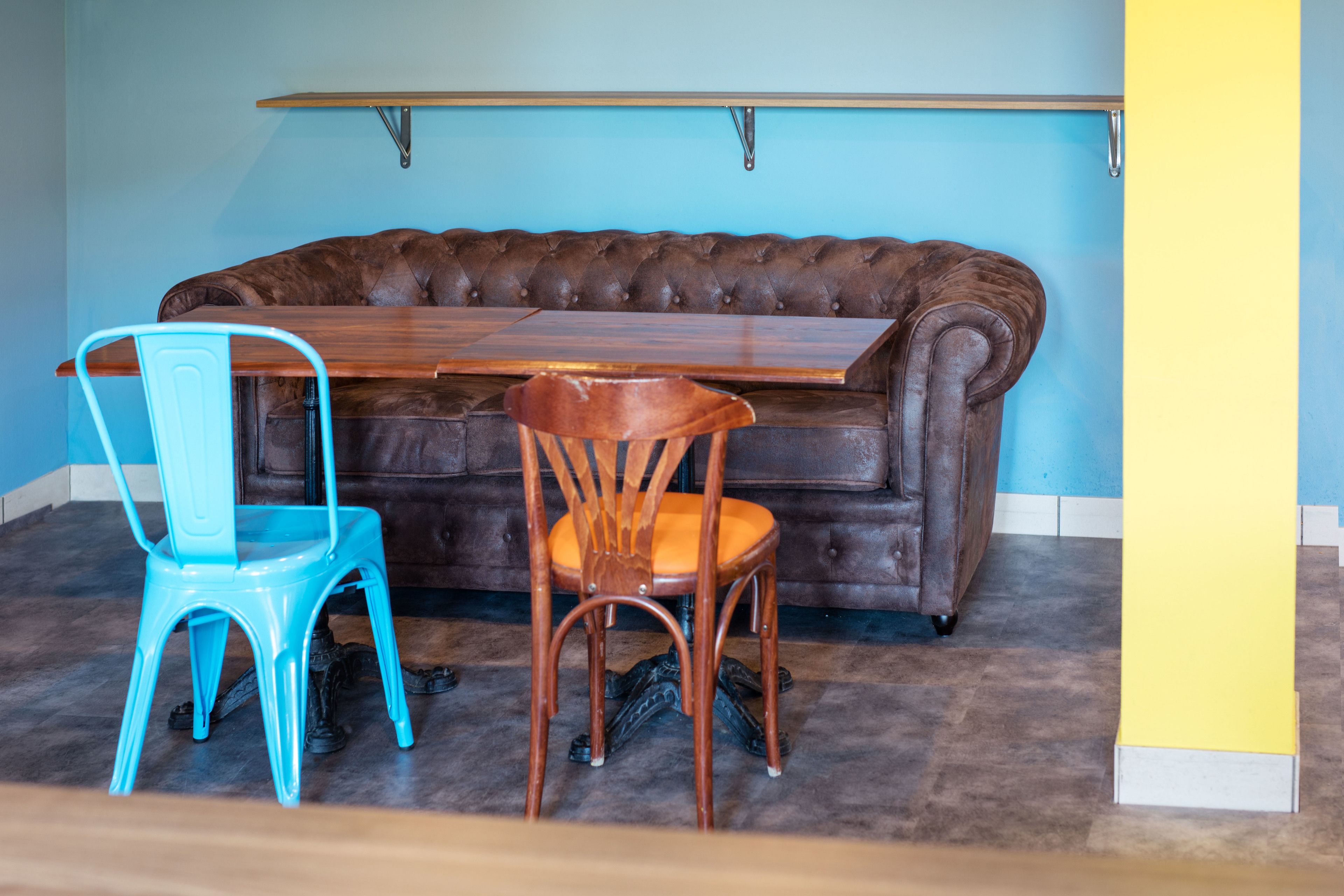 A room with a brown leather sofa, a wooden table, and two chairs, one blue and one brown.