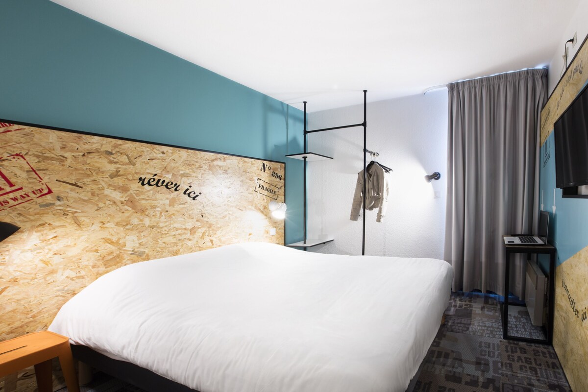A modern hotel room with a large bed, a wooden headboard with text, a small desk with a laptop, a hanging coat rack, and a TV mounted on the wall.