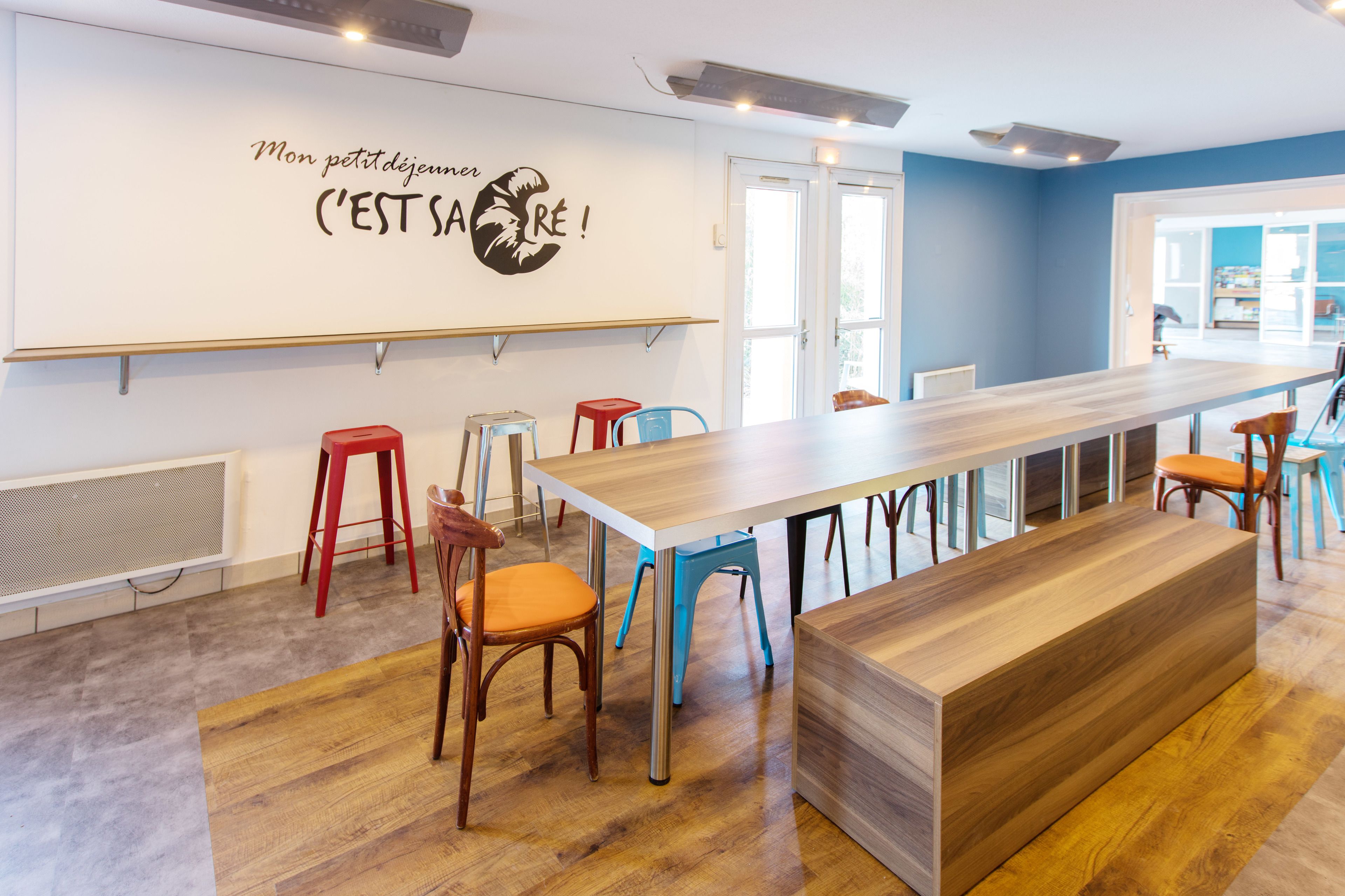 A modern dining area with a long wooden table, red and blue chairs, and a bench.