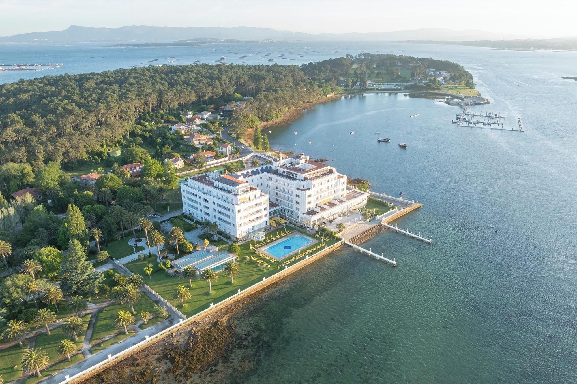 An aerial view of a coastal hotel with a large building, a swimming pool, and a pier extending into the water.