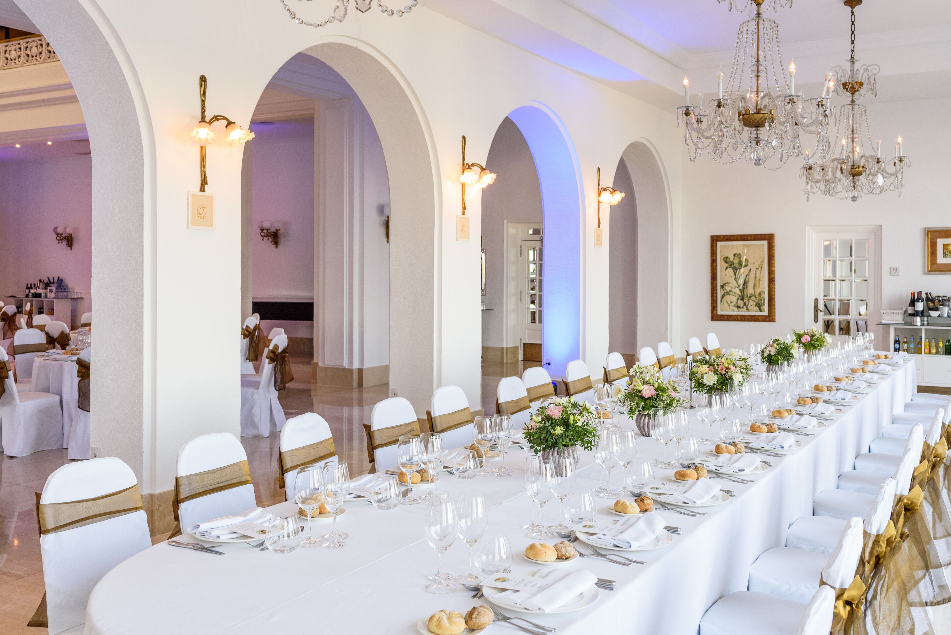 Una larga mesa de banquete decorada con manteles blancos, fajines dorados y centros de mesa florales.