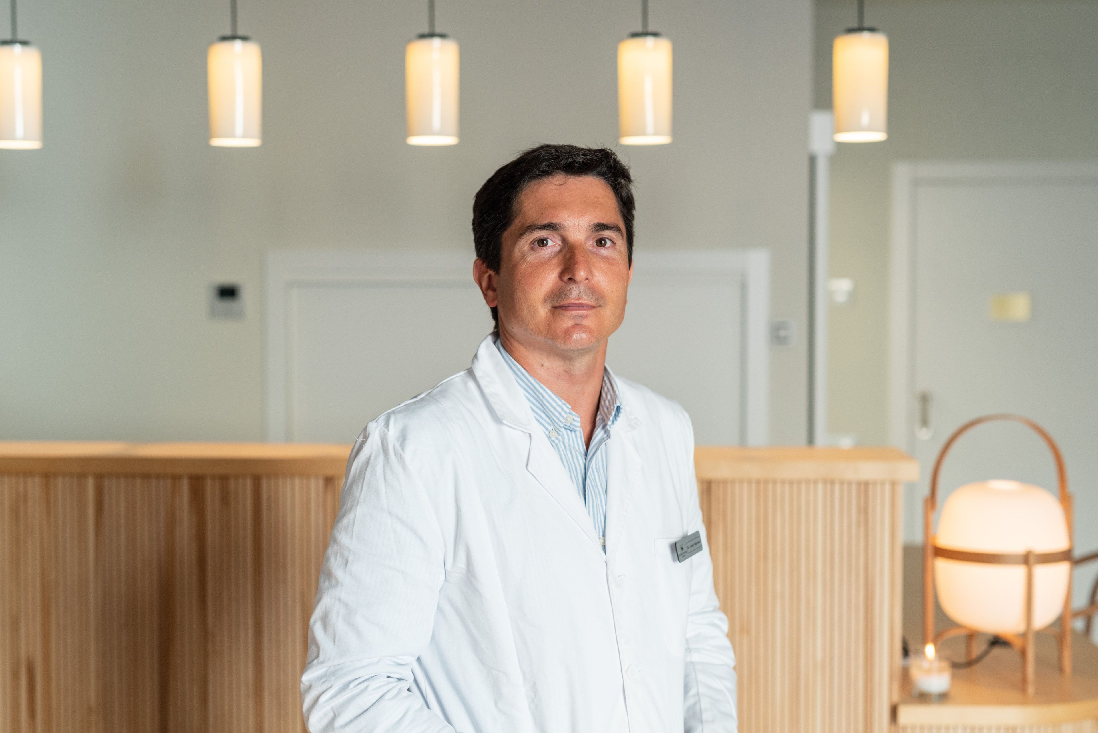 A person in a white lab coat standing in a room with wooden paneling and pendant lighting.