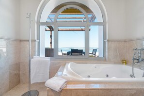 Een badkamer met een groot bad, een douchecabine van glas en uitzicht op zee door een rondbogig raam.