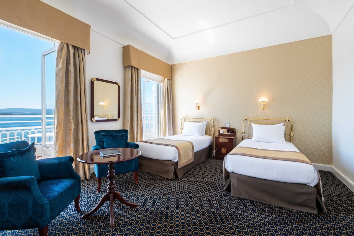A hotel room with two beds, a sitting area with a table and chairs, a large window with curtains, and a view of the sea.