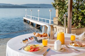 A breakfast table with croissants, fruit, and beverages by a lakeside view.