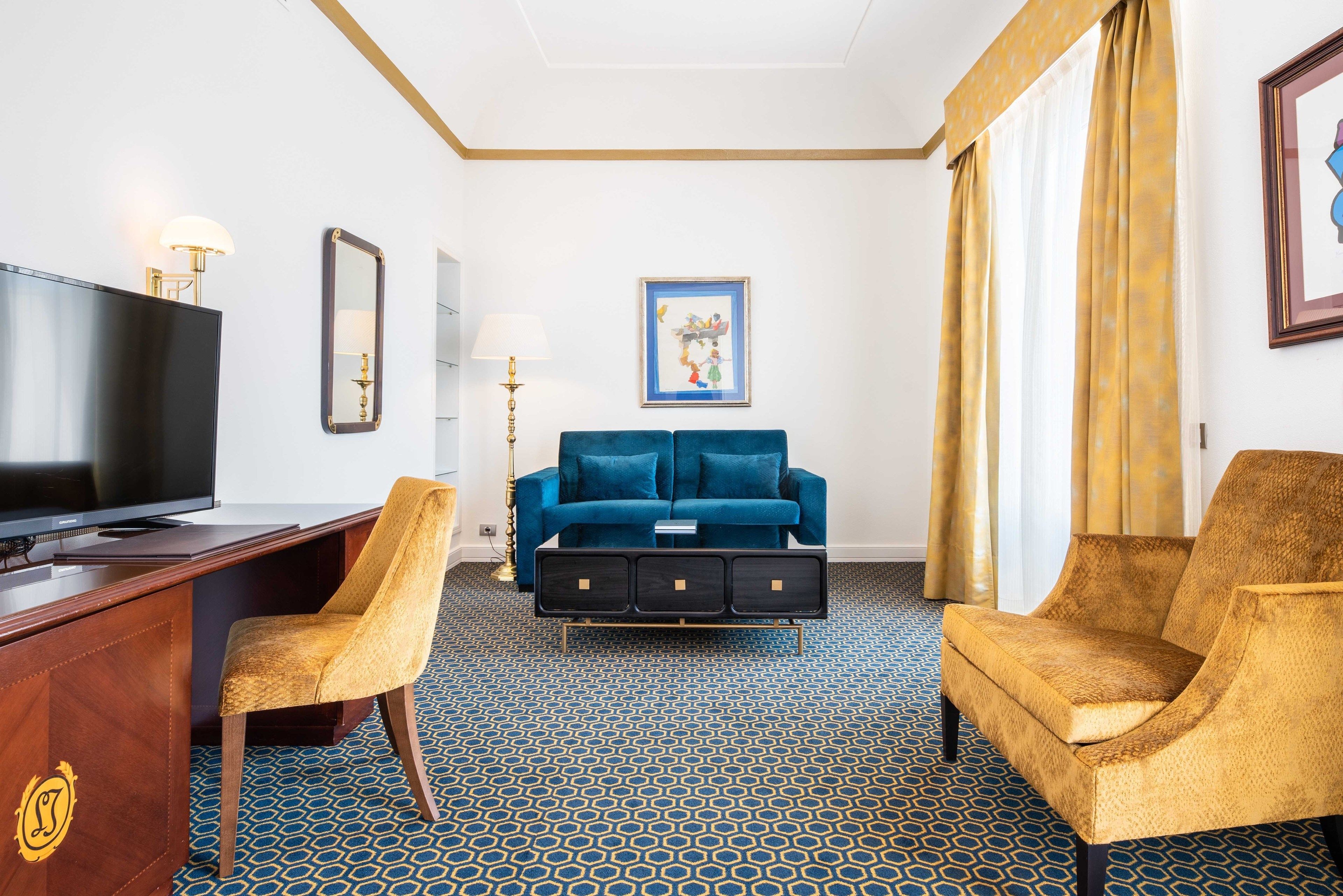 Habitación de hotel con televisión, dos sillones, un sofá, un escritorio y alfombra estampada.