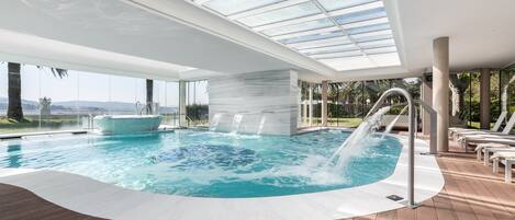 Piscina interior moderna com teto de vidro, cercada por espreguiçadeiras e um deck de madeira.