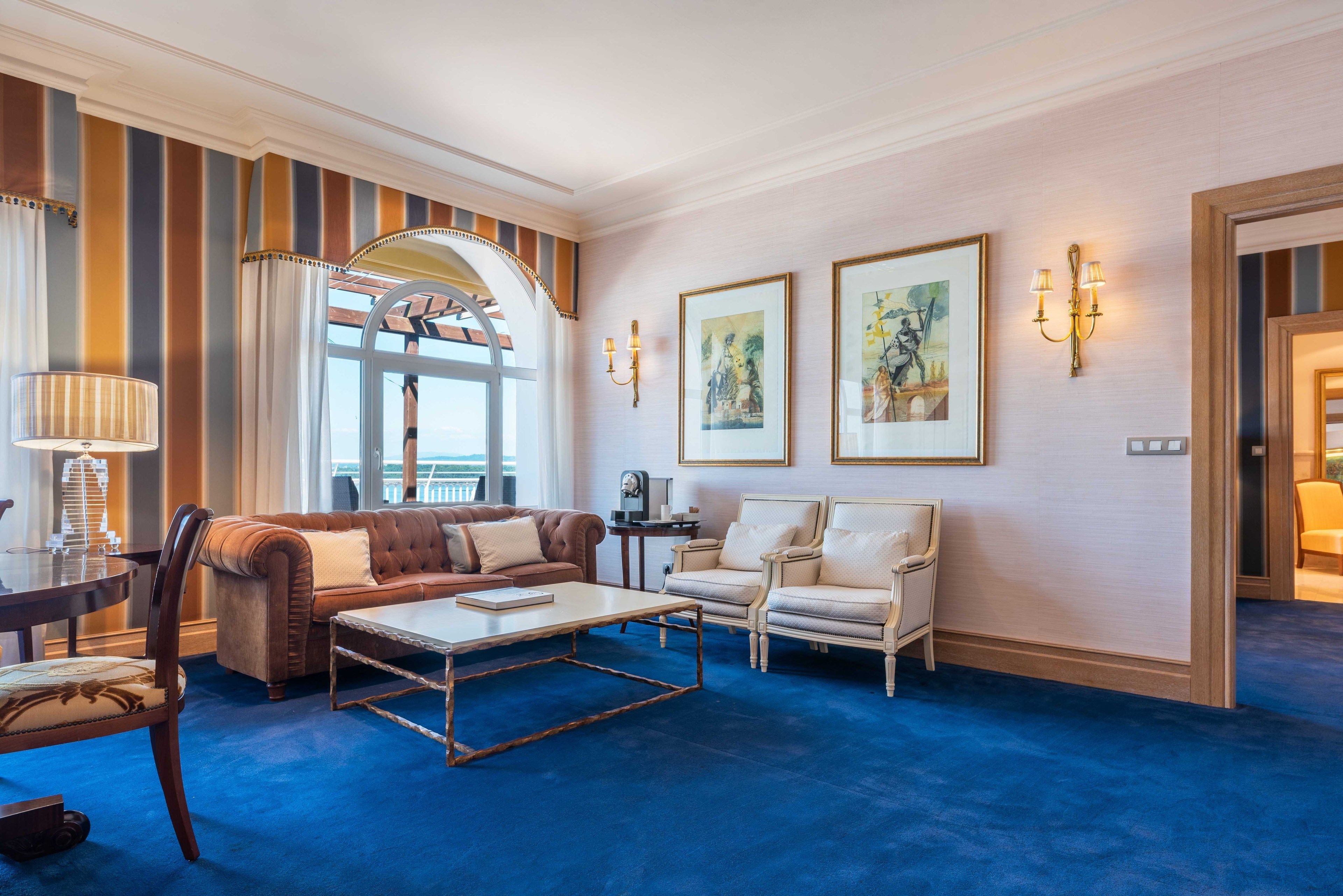 Un salón amplio con alfombra azul, sofá marrón, una mesa de centro blanca y dos sillones blancos. Se observan obras de arte enmarcadas en la pared y un ventanal con cortinas.