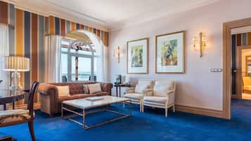 Un salón amplio con alfombra azul, sofá marrón, una mesa de centro blanca y dos sillones blancos. Se observan obras de arte enmarcadas en la pared y un ventanal con cortinas.