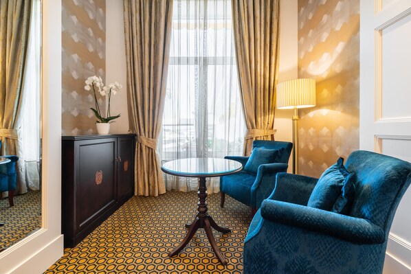 A room with a patterned floor, a round table, two blue armchairs, a dark wooden cabinet, a potted orchid, and a lamp.