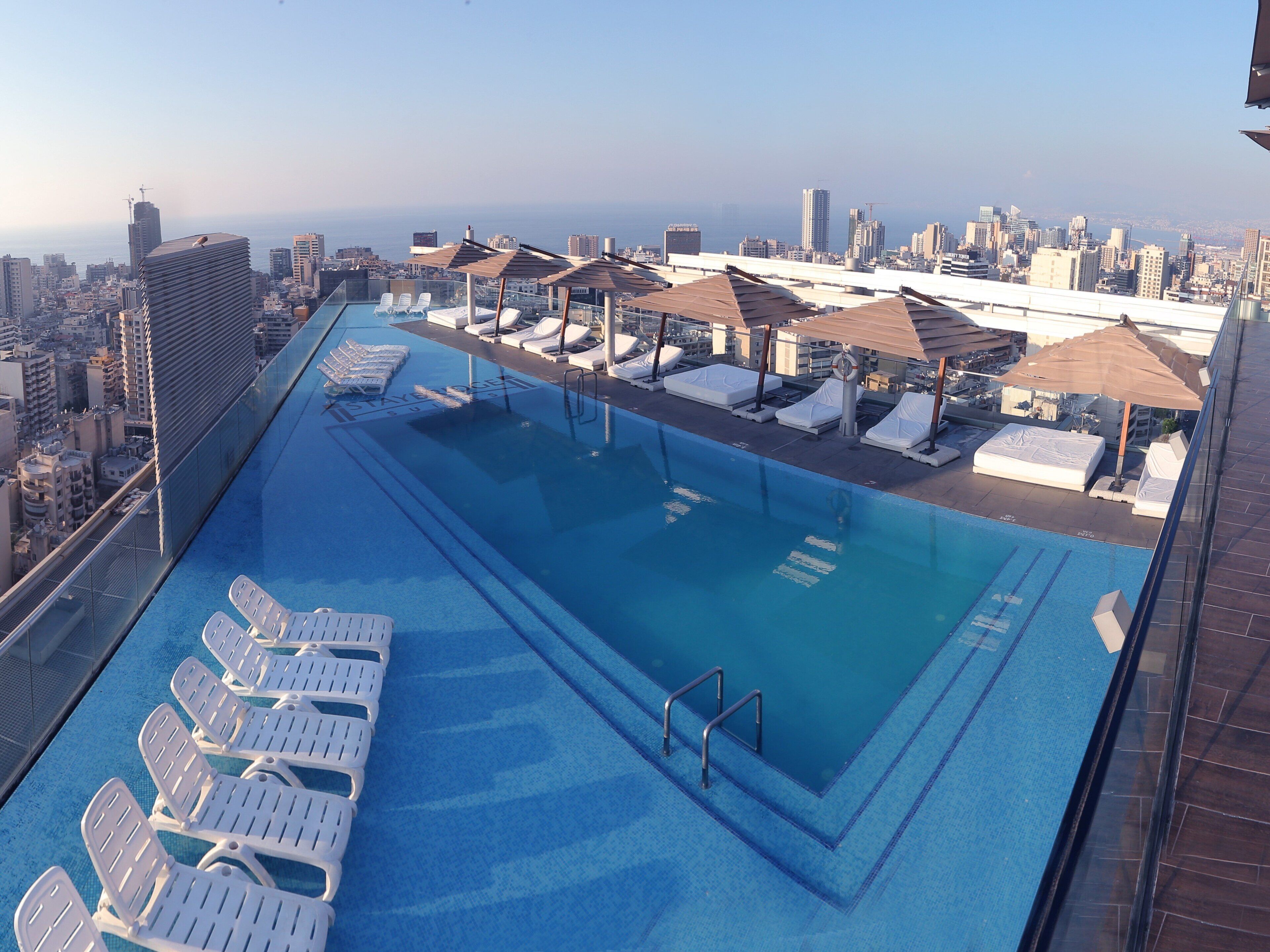A rooftop pool with sun loungers and umbrellas, overlooking a cityscape.