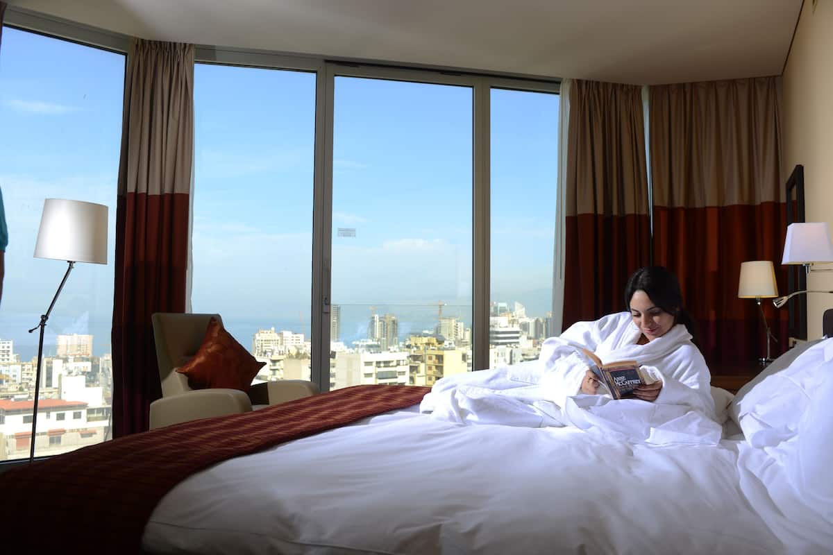 A hotel room with a large bed, a chair, a lamp, and a view of the cityscape through large windows.