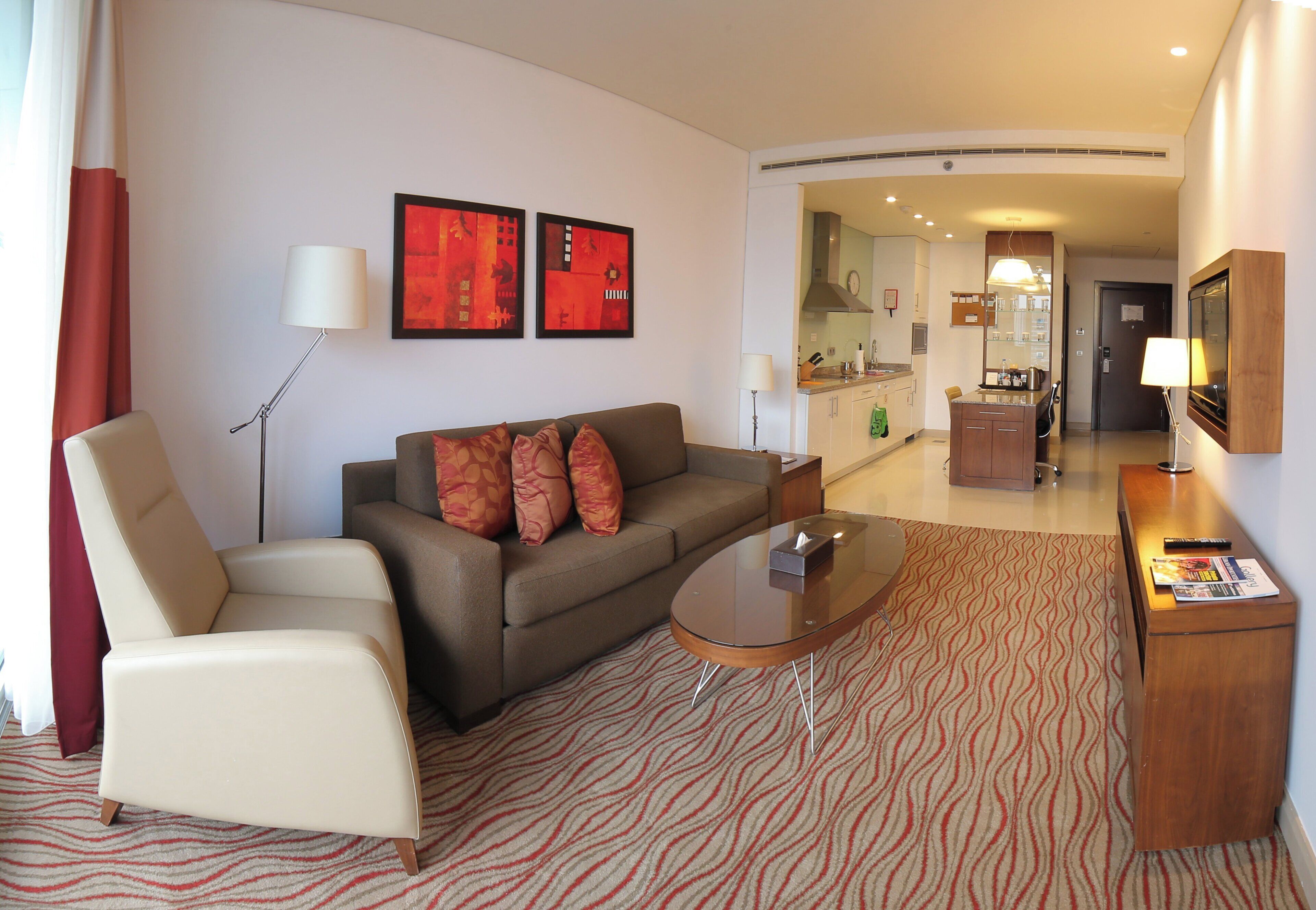 A hotel room with a sofa, armchair, a small coffee table, a TV, and a kitchen area visible through an open door.