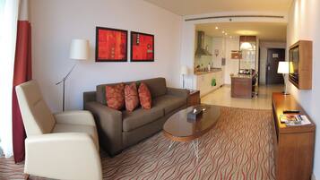 A hotel room with a sofa, armchair, a small coffee table, a TV, and a kitchen area visible through an open door.