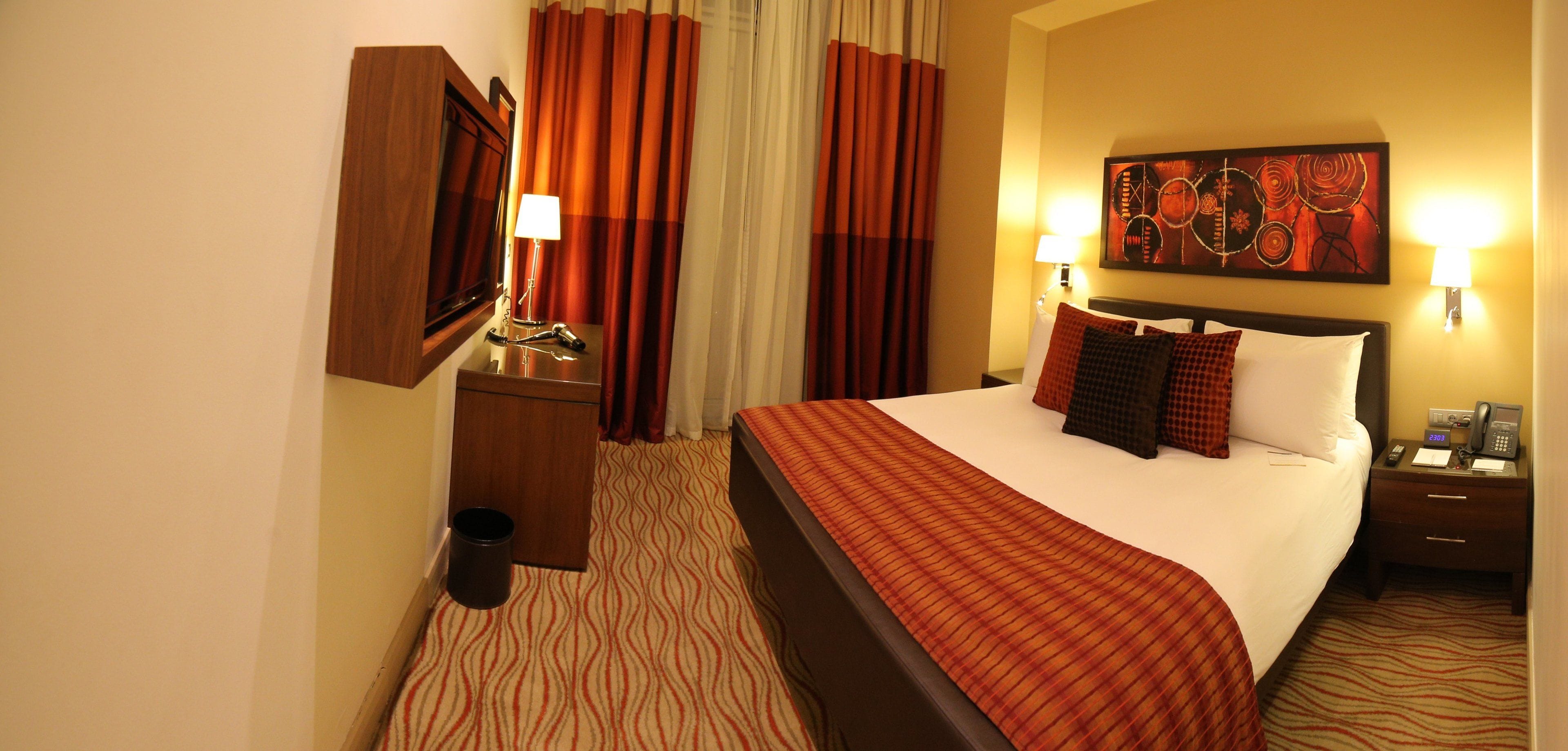 A hotel room with a bed, a nightstand with a lamp, a telephone, and a painting on the wall.