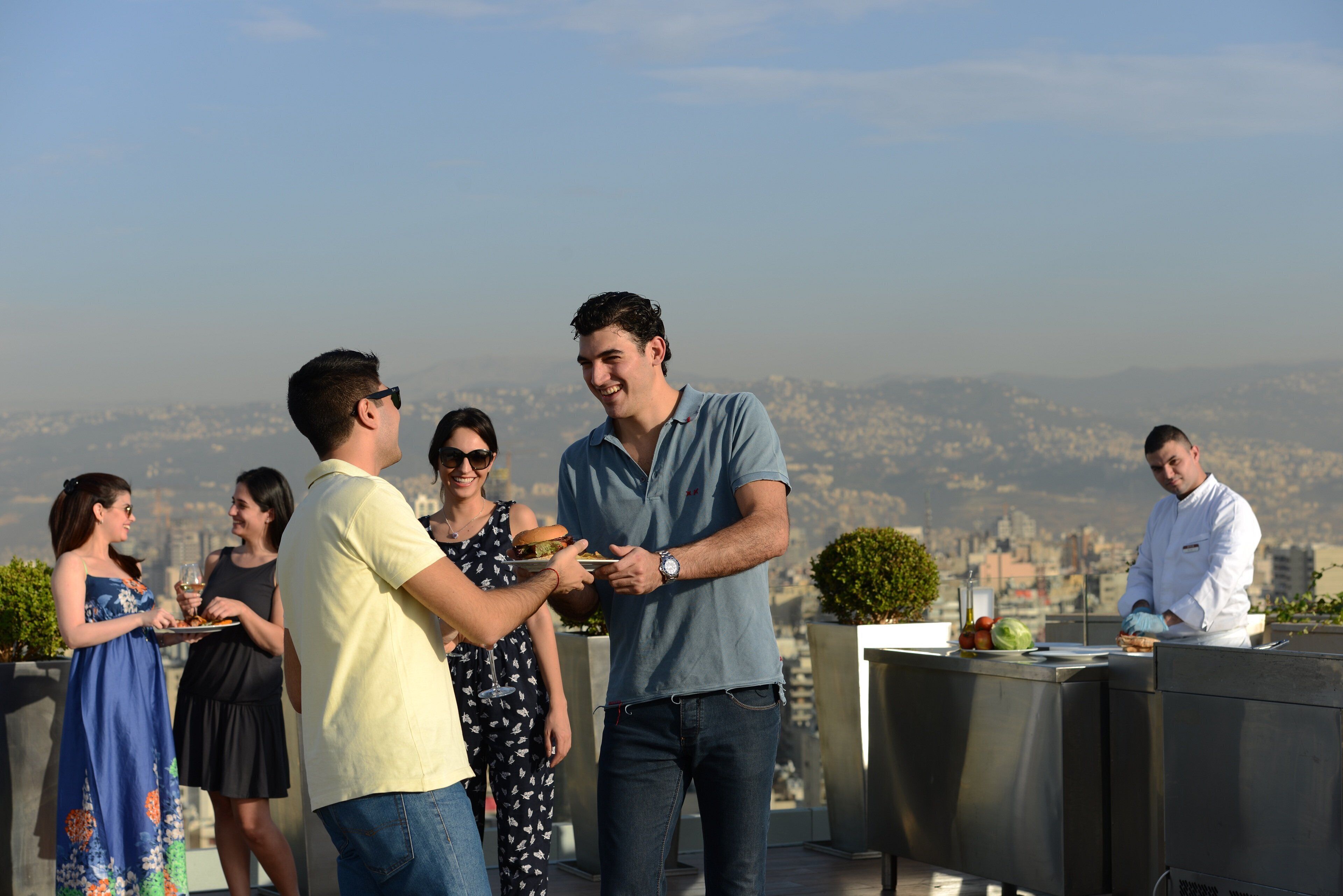 A rooftop terrace with a city view, outdoor seating, and a chef preparing food.