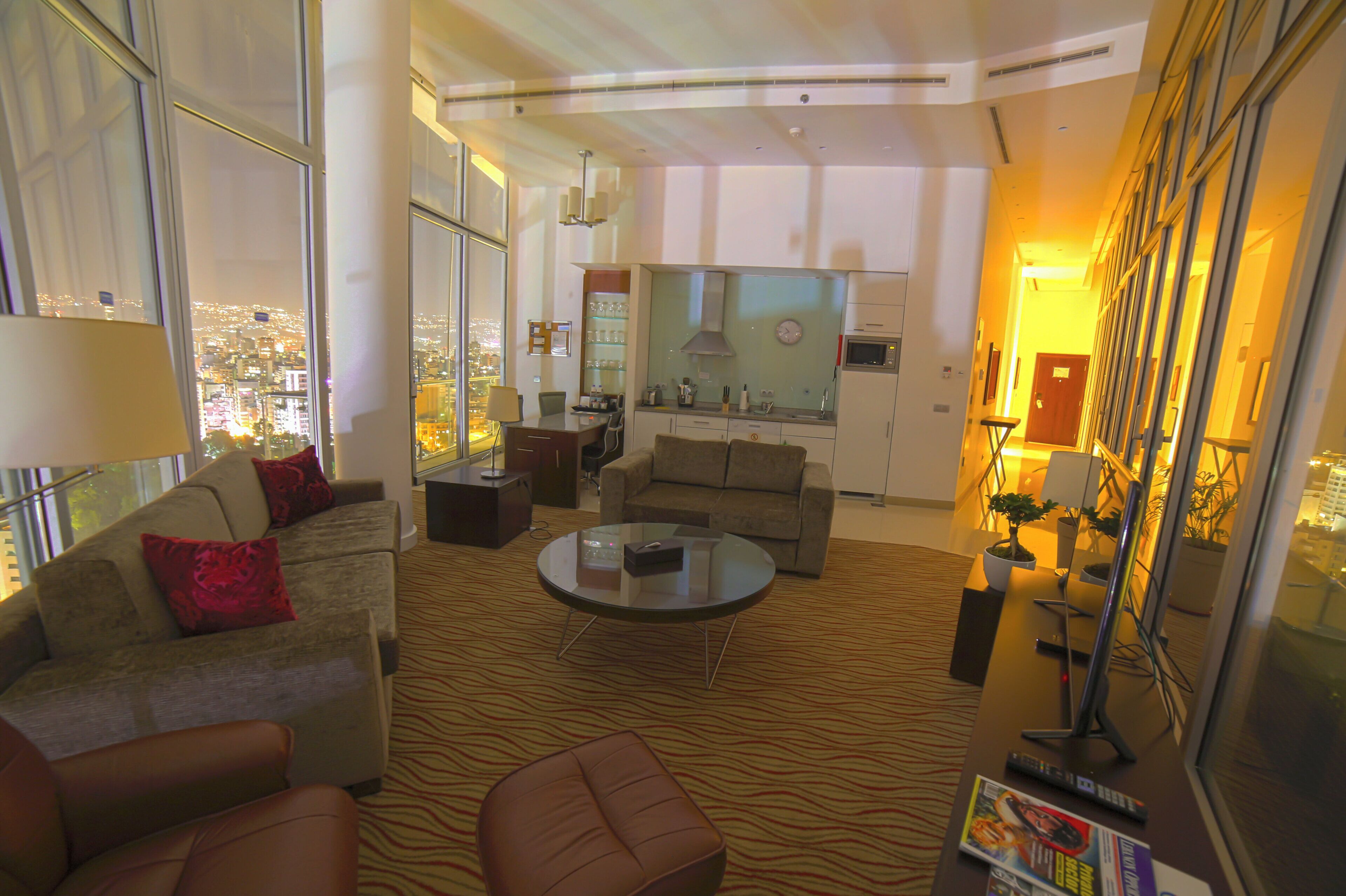 A modern hotel room with a city view, a living area with a sofa and armchairs, a dining area with a kitchen, and a large window.