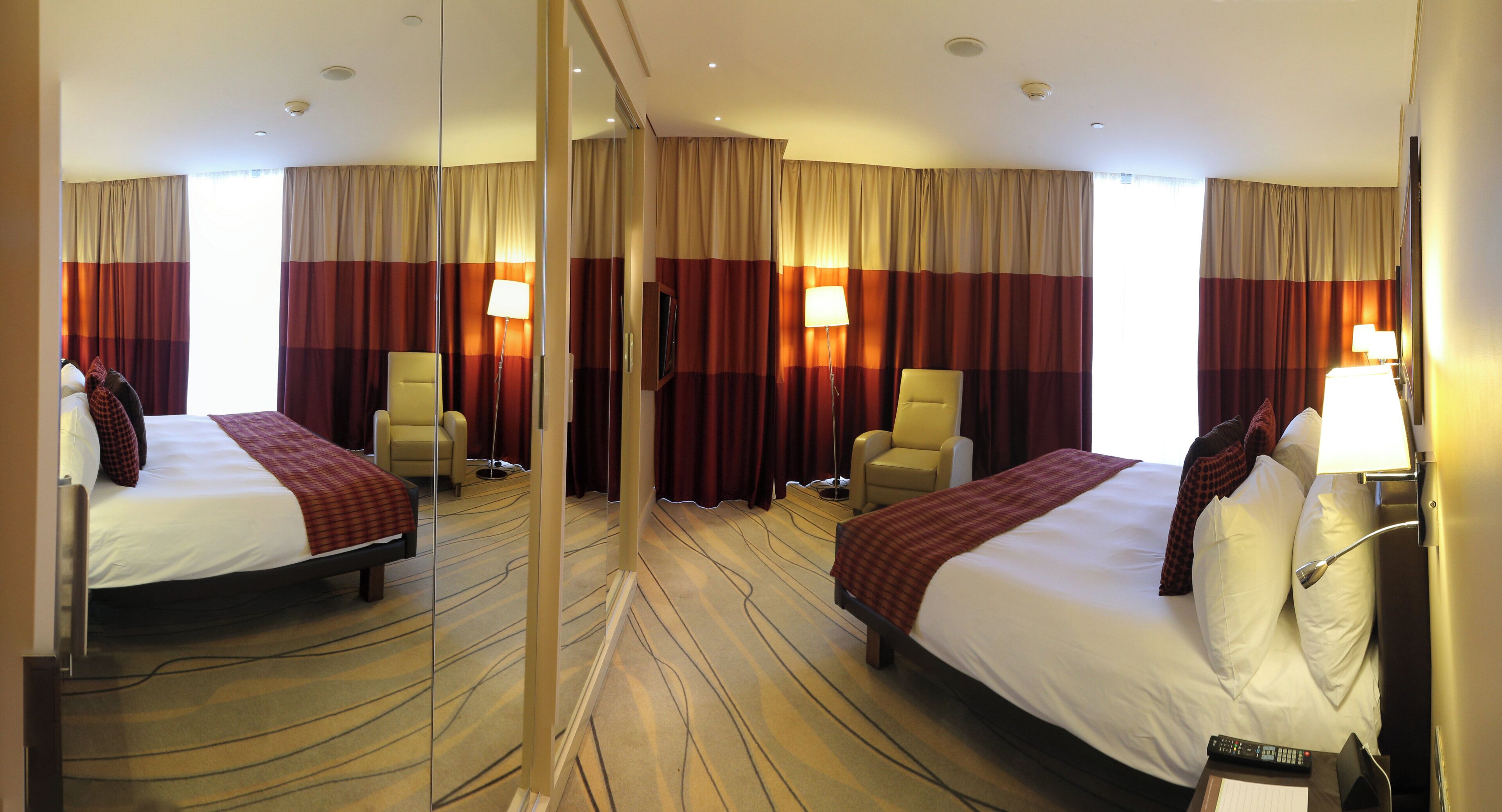 A hotel room with two beds, a mirror, a chair, a lamp, and a remote control on a side table.