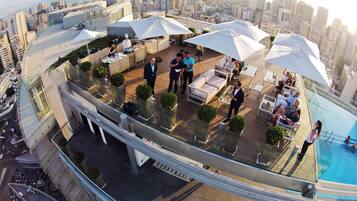 A rooftop bar with outdoor seating and a pool, overlooking a cityscape.