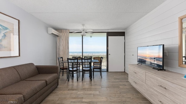 A room with a sofa, dining table with chairs, a TV on a wooden cabinet, and a view of the beach through a glass door. - Beau Rivage Beach Resort (Wildwood Crest)