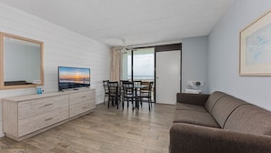 A modern living room with a flat-screen TV, a wooden cabinet, a brown sofa, and a dining area with chairs. - Beau Rivage Beach Resort (Wildwood Crest)