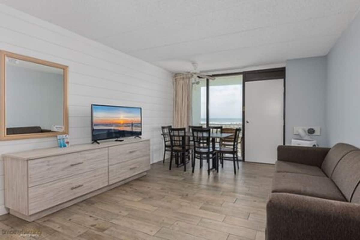 A modern living room with a flat-screen TV, a wooden cabinet, a brown sofa, and a dining area with chairs.