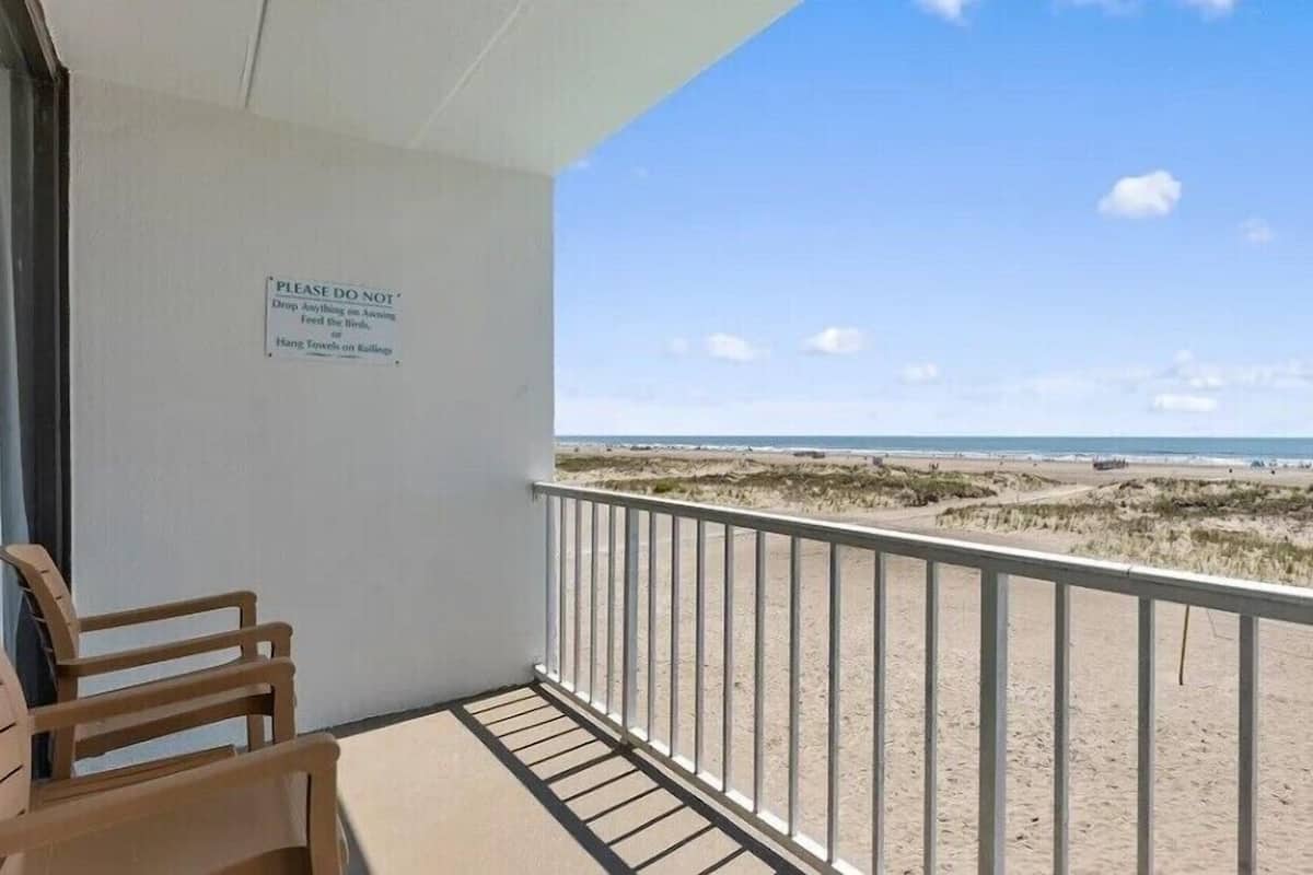 A balcony with a view of the beach, a sign advising against hanging towels on railings, and two wooden chairs.