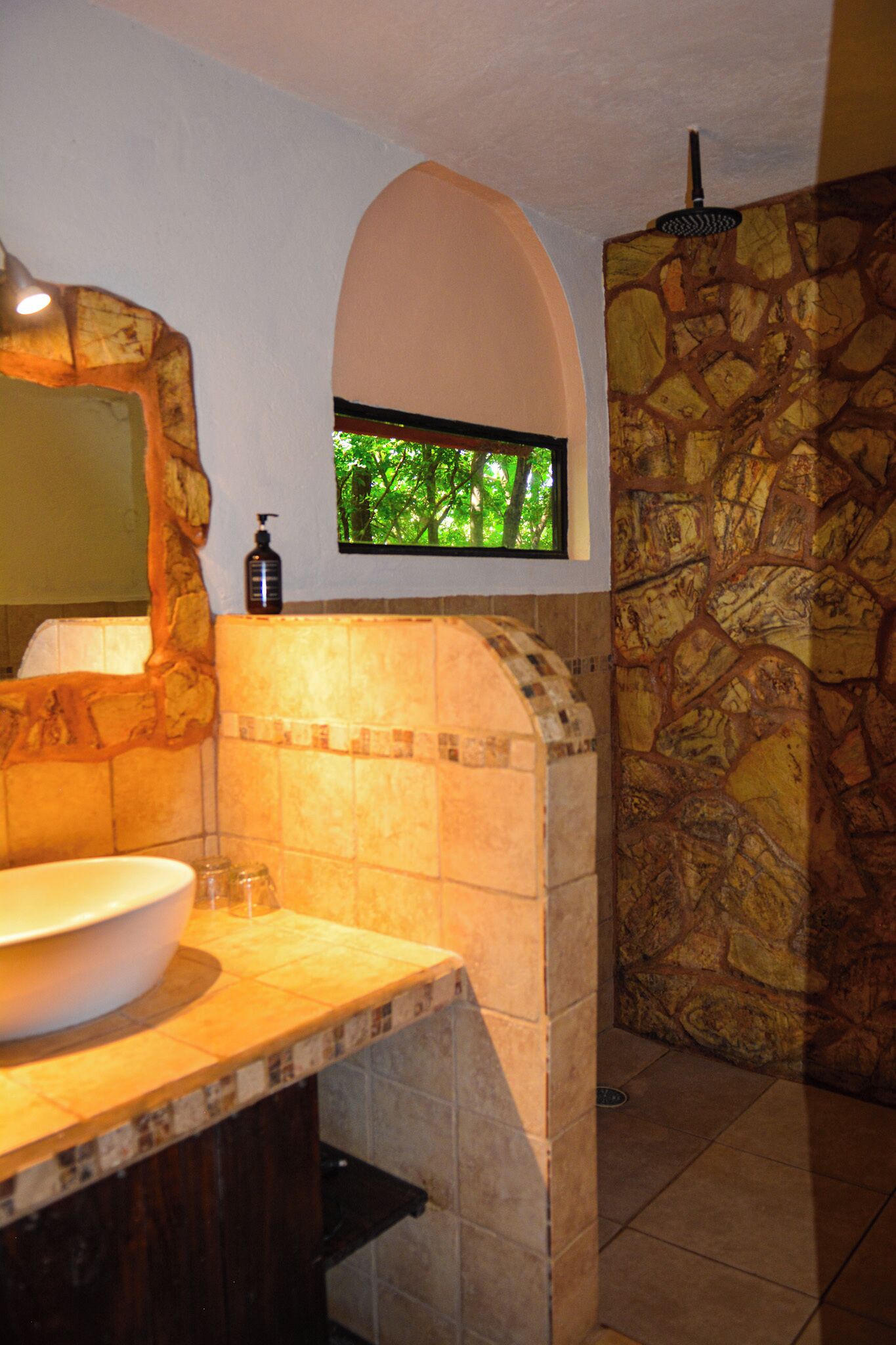 A bathroom with a stone wall, a sink with a marble countertop, a window with a view of trees, and a mirror with a stone arch above it.