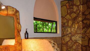 A bathroom with a stone wall, a sink with a marble countertop, a window with a view of trees, and a mirror with a stone arch above it.