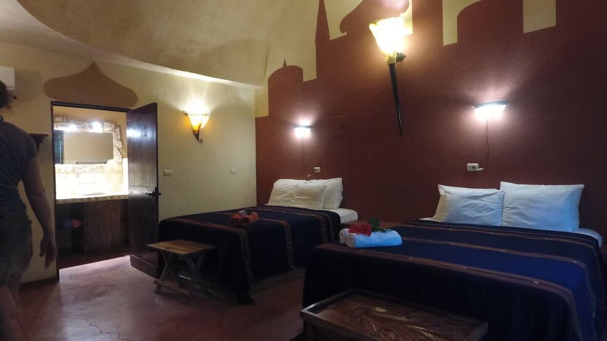 A hotel room with two beds, a small table, and a bench. There are wall-mounted lights and a door leading to a bathroom.