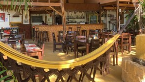 Un espace de restauration extérieur accueillant, avec des tables et des chaises en bois, un hamac et un bar où sont exposées des bouteilles, ainsi qu’un tableau noir servant de menu.