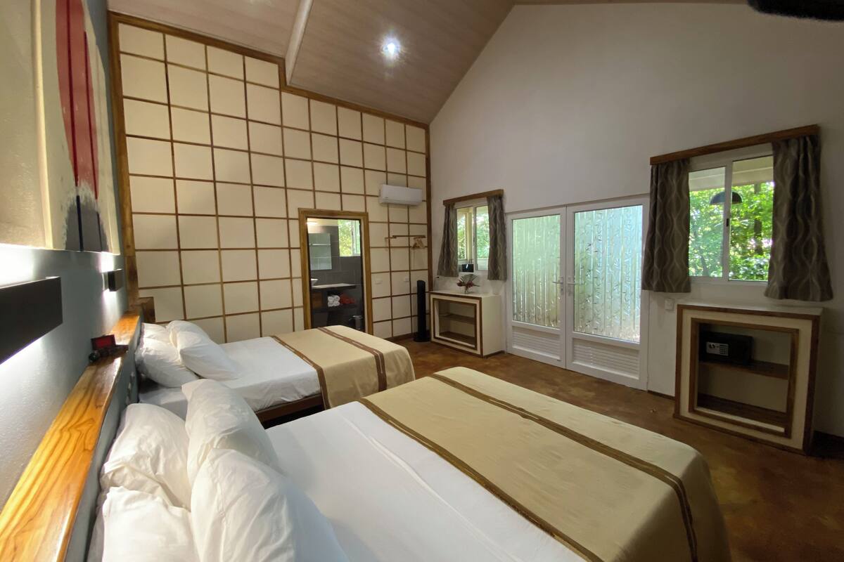 Hotelkamer met drie bedden, een houten bureau, een spiegel, een raam met gordijnen en uitzicht op groen.