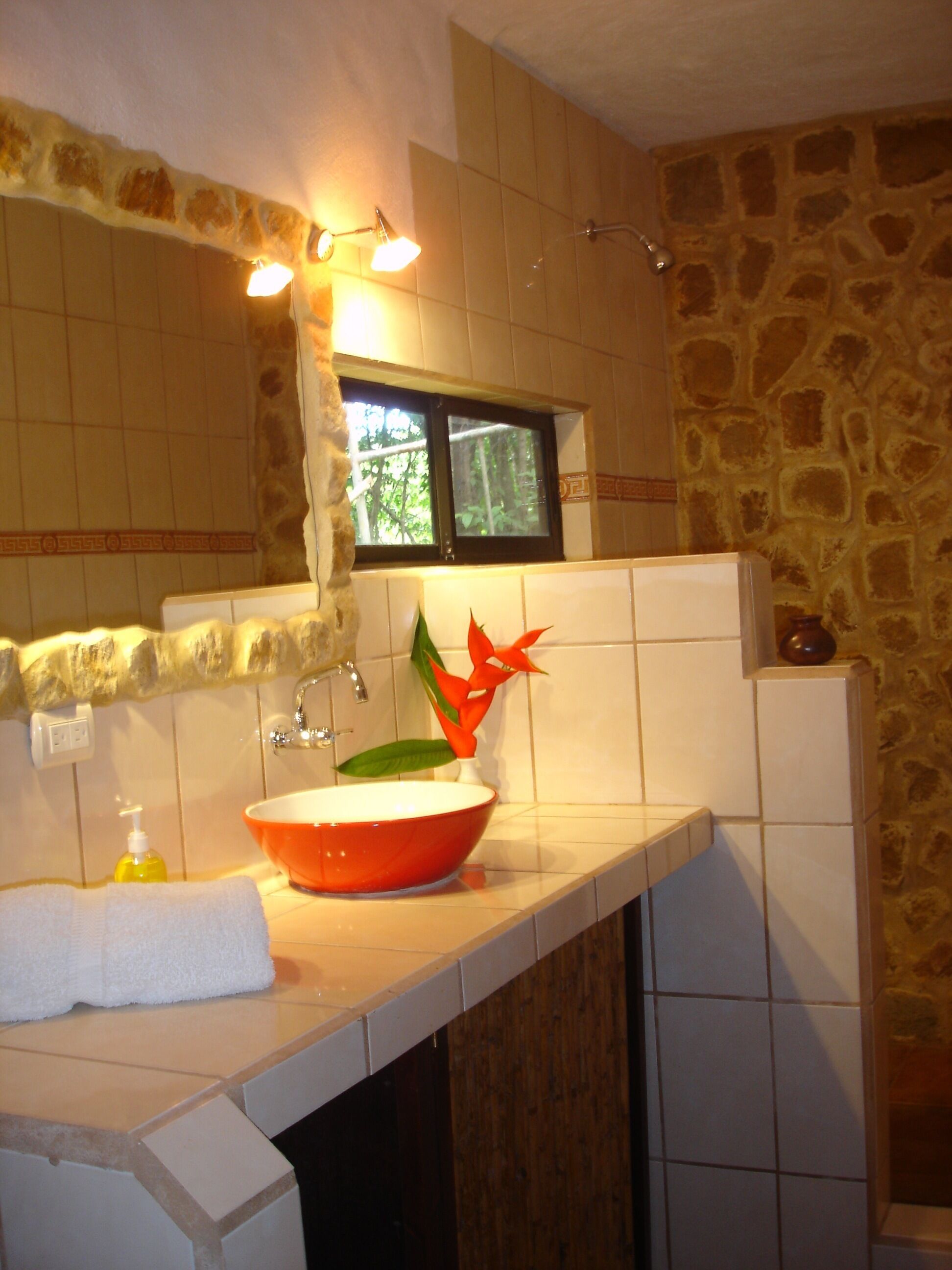 Een badkamer met een stenen muur, een witte wastafel met een rode kom, een spiegel, een raam en een handdoek op het aanrecht.