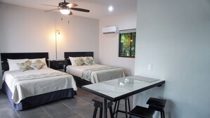 Habitación de hotel con dos camas, ventilador de techo, aire acondicionado, ventana con vistas y una pequeña mesa con taburetes.
