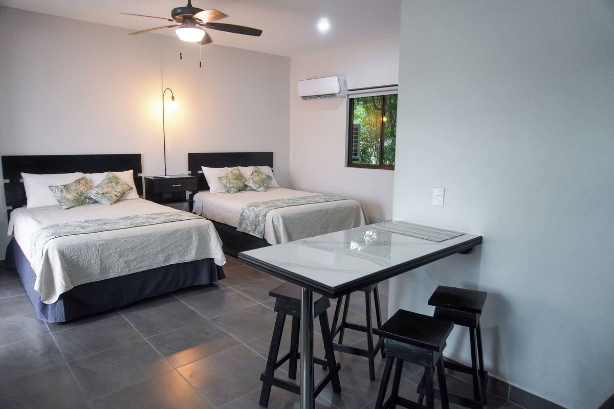 Habitación de hotel con dos camas, ventilador de techo, aire acondicionado, ventana con vistas y una pequeña mesa con taburetes.