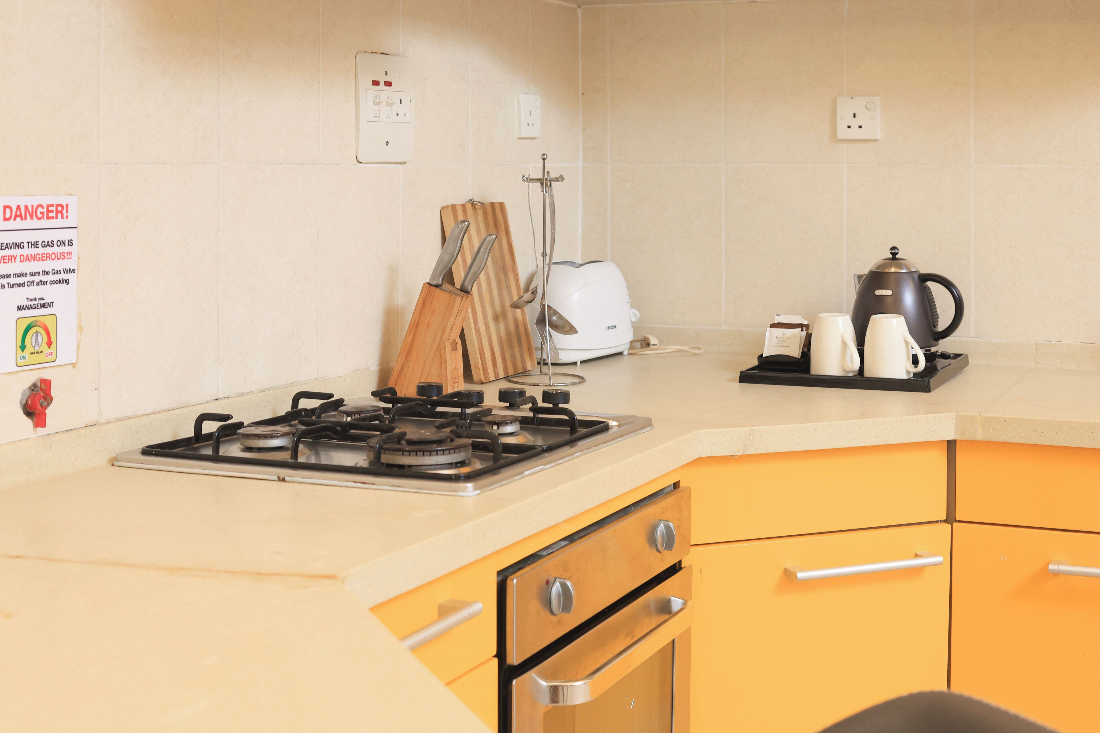 Una cocina con estufa a gas, tostadora, hervidor de agua y cafetera sobre el mostrador. Hay cuchillos y una tabla de cortar en la pared.