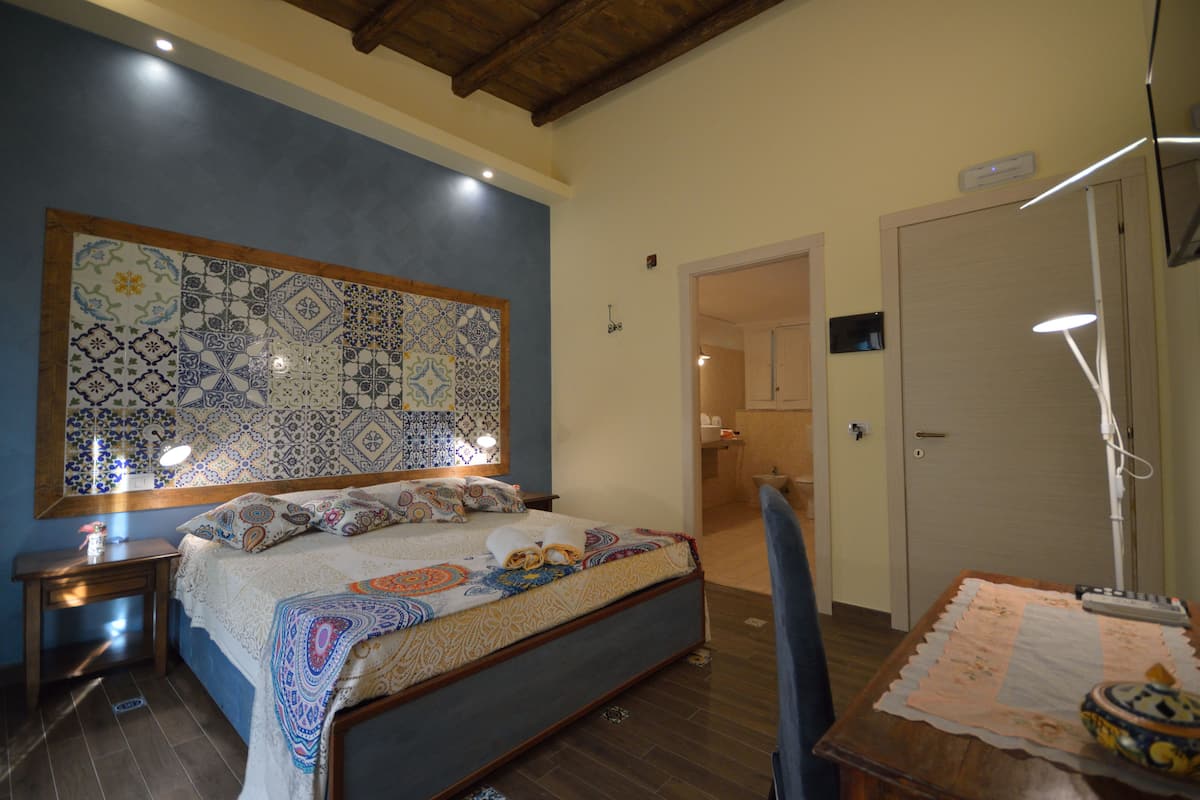 A bedroom with a large bed, bedside tables, a desk with a lamp, and a bathroom visible through an open door.