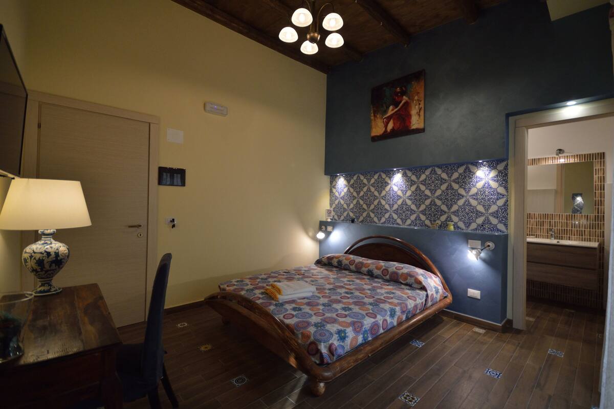 A bedroom with a bed, a chair, a lamp, a door, and a bathroom visible through an open door.