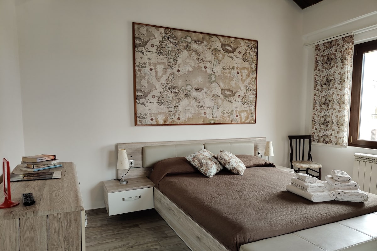 A bedroom with a large bed, a bedside table with books, a lamp, and a map on the wall.