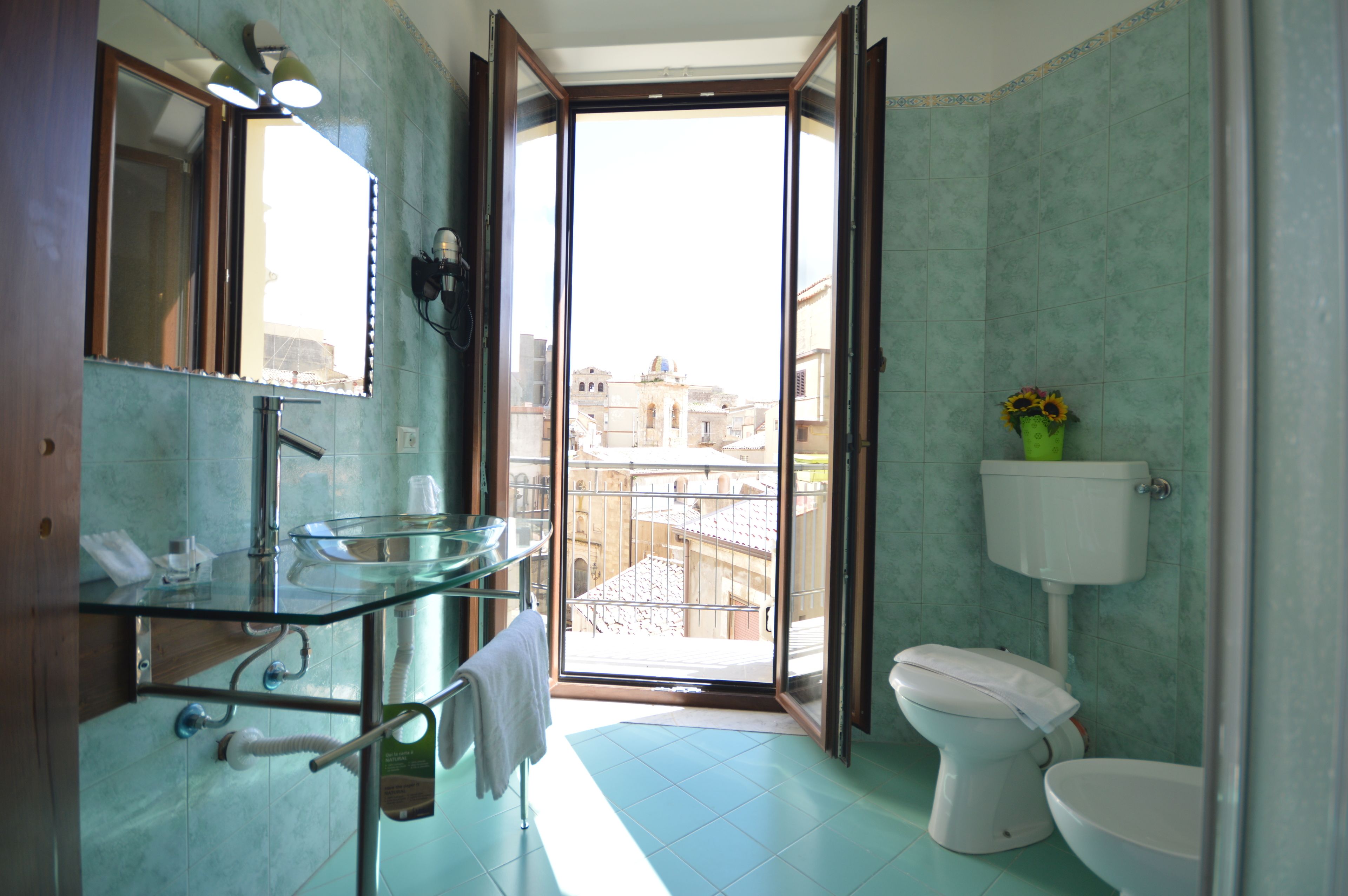 Ein Badezimmer mit einem Waschbecken aus Glas, einer Toilette und Blick auf Gebäude durch eine offene Tür.