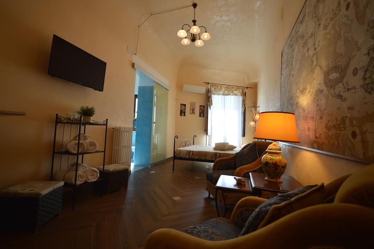 A cozy living room with a sofa, a table with a lamp, a TV mounted on the wall, and a bed with a window.