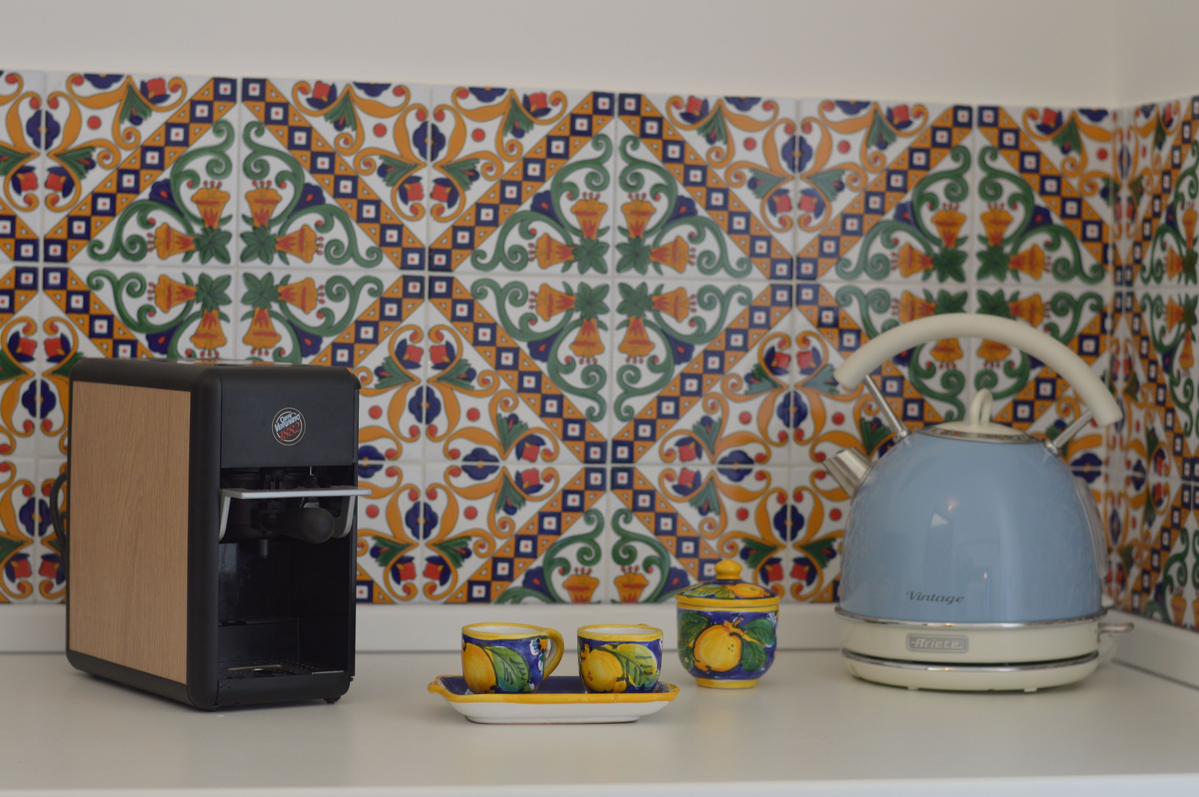 Una cafetera, dos tazas y una tetera sobre una encimera con un salpicadero de baldosas estampadas.