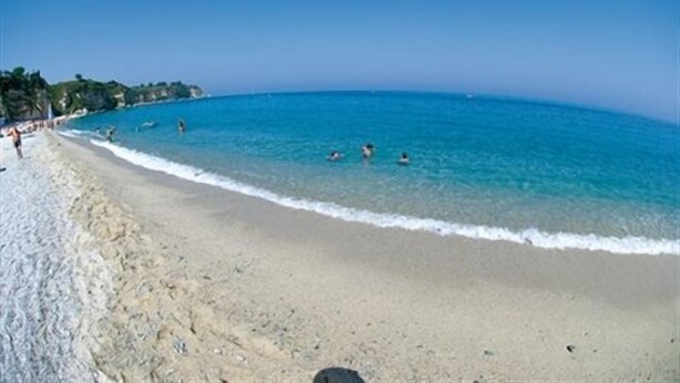 Une plage avec une eau d’un bleu limpide, un rivage sablonneux et quelques personnes qui se baignent.