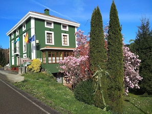 A green building with a sign indicating it is a hotel and restaurant. There are flowering trees and a well-maintained lawn in front. - Casa Vieja del Sastre (Cudillero)