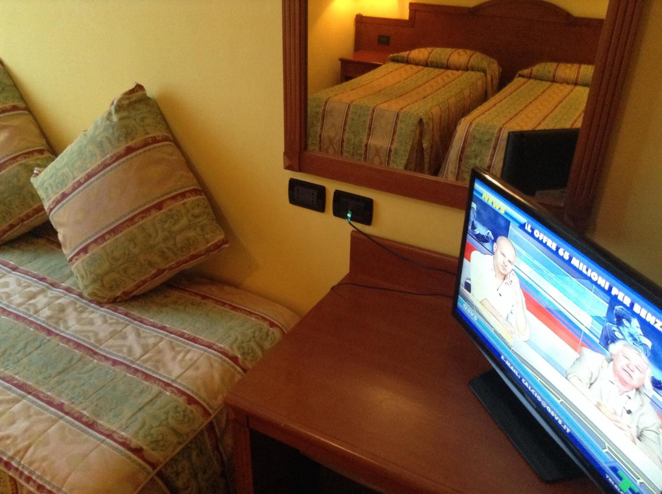 Camera d'albergo con due letti, una TV su un tavolino di legno e una presa di corrente a parete.