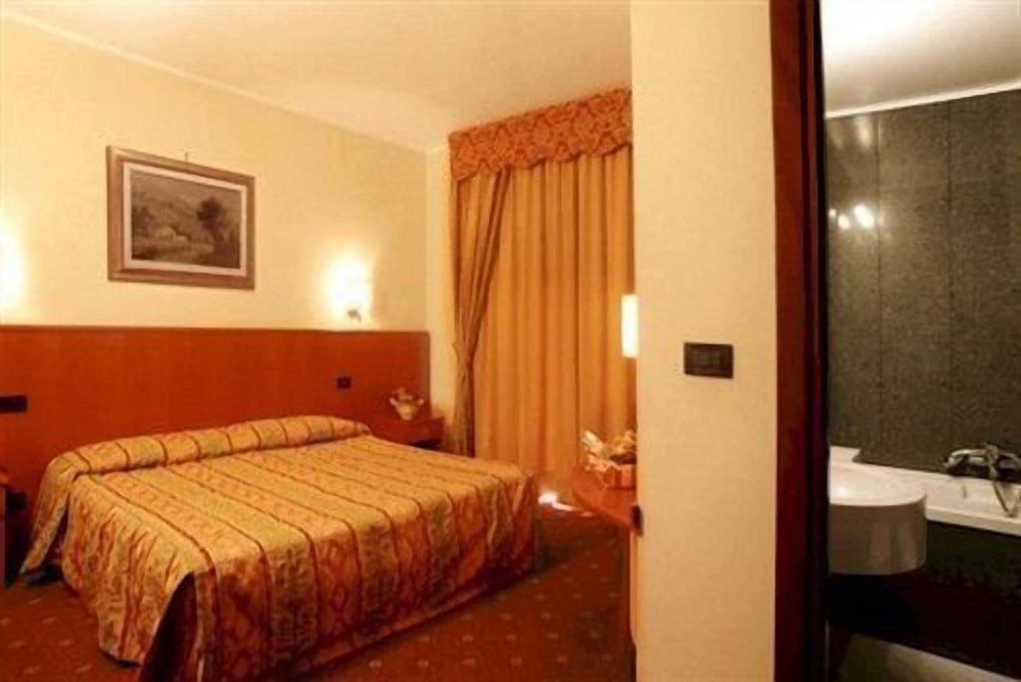 Habitación de hotel con cama doble, mesita de noche, lámpara, un cuadro en la pared y baño visible a través de una puerta abierta.