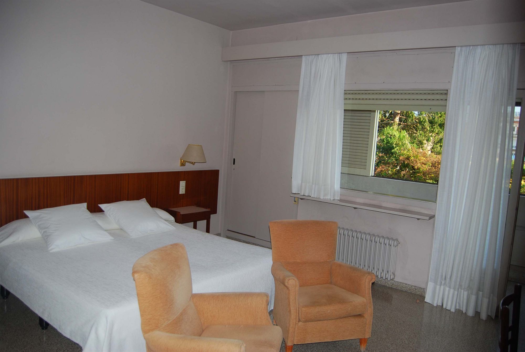 Habitación de hotel con cama, dos sillones, una mesita de noche con lámpara, ventana con cortinas y un radiador.