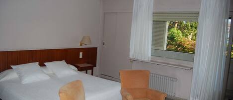 Habitación de hotel con cama, dos sillones, una mesita de noche con lámpara, ventana con cortinas y un radiador.