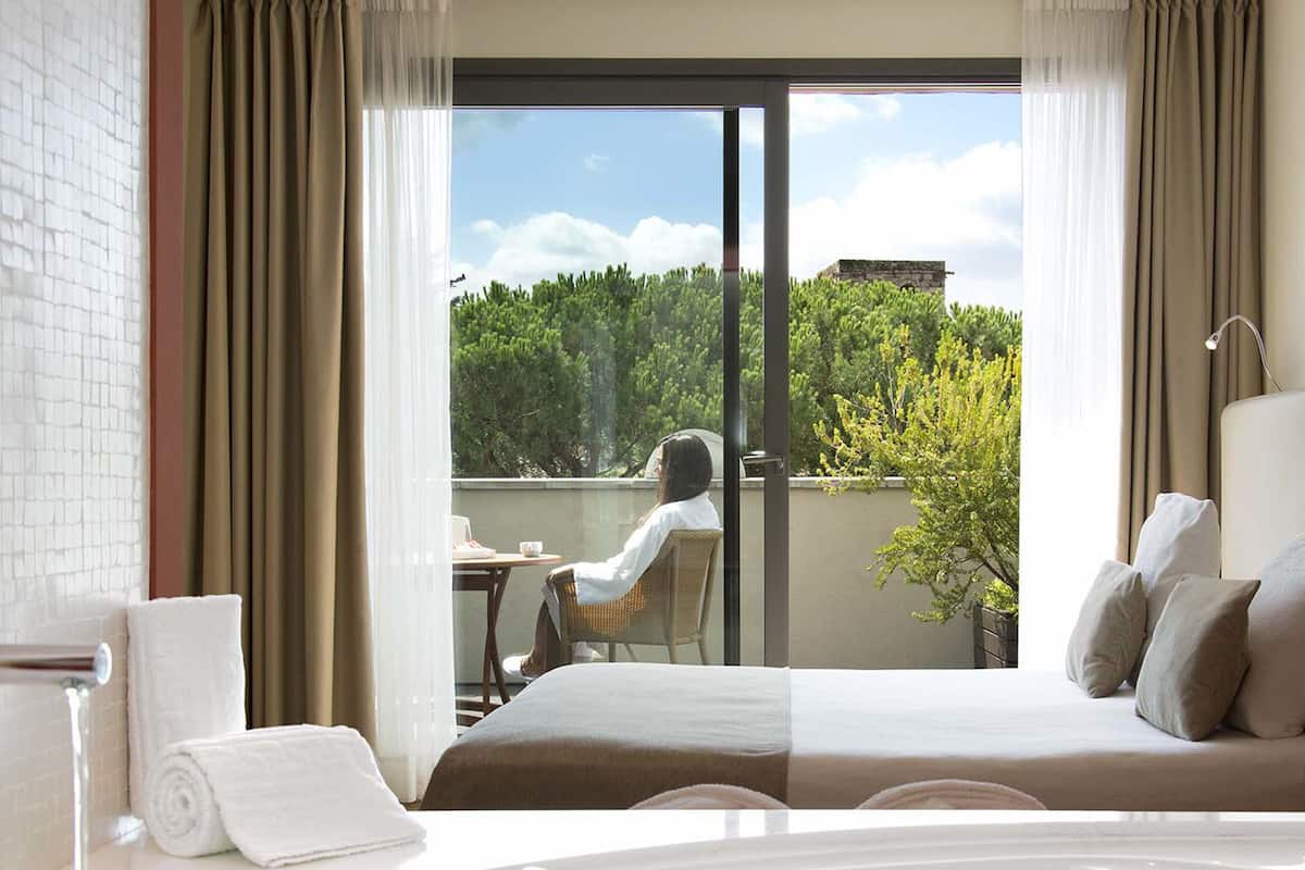 A hotel room with a large bathtub, a balcony with a view of greenery, and a bed with pillows.